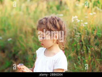 Schönes kleines Mädchen, das mit Blumen im sonnigen Frühlingspark spielt. Fröhliches süßes Kind, das bei Sonnenuntergang Spaß im Freien hat. Stockfoto