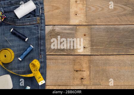 Hose aus blauem Denim-Baumwollstoff mit Schneiderwerkzeugen auf braunem Holzhintergrund - Jeansmode Mending oder Reparaturkonzept, flacher Lay-Draufsicht von oben Stockfoto