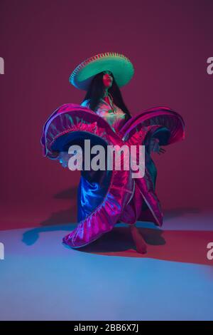 Fabelhafte Cinco de Mayo Tänzerin im violetten Studio Hintergrund in Neonlicht. Wunderschönes weibliches Modell in traditionellem Kostüm und Sombrero Tanz. Celebration, Urlaub, Schönheit und Modekonzept. Stockfoto