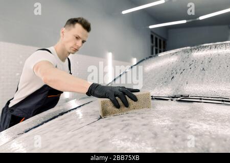 Autowaschkonzept. Junger konzentrierter kaukasischer männlicher Arbeiter, der den Schwamm in der Hand hält und die Autohaube mit Seife säubert. Selektiver Fokus auf der Hand. Stockfoto