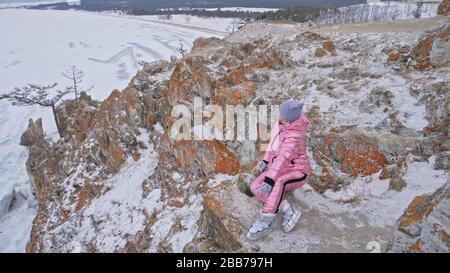 Frau sitzt auf dem Hügel. Der Reisende geht in den oberen Bergen. Das schöne Mädchen ist mit einer modischen pinkfarbenen Daunenjacke bekleidet. Explorer sieht in den Himmel. Stockfoto