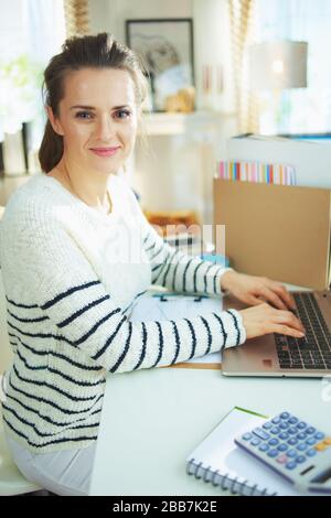 Fröhliche, moderne 40 Jahre alte Geschäftsfrau im gestreiften Pullover im provisorischen Heimbüro im modernen Haus bei sonniger Tagesarbeit. Stockfoto
