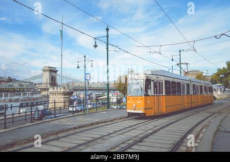 Budapest, Ungarn - 6. November 2019: Gelbe Straßenbahn an einer Straßenbahnstation. Szechenyi-Kettenbrücke und das historische Zentrum im Hintergrund. Öffentliche Verkehrsmittel in Ungarn. Horizontales Foto mit Filter. Stockfoto