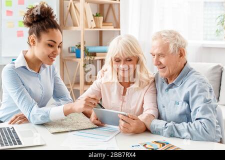 Reisebüro im Büro, das älteren Kunden den Reiseplan auf einem digitalen Tablet zeigt Stockfoto