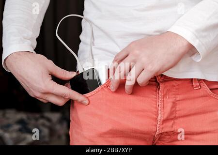 Mit der männlichen Hand wird die Powerbank und das Mobiltelefon in die vordere Tasche der korallenfarbenen Jeans geladen. Stockfoto