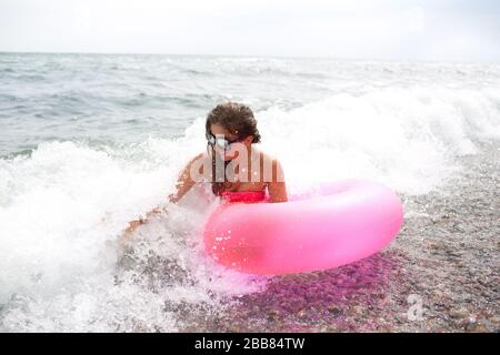 Fröhliche junge Frau sitzt an der Küste oder am Meer in Wellen mit rosafarbener Luftmatratze in der Hand und genießt. Urlaubs- oder Reisezeit. Schaum aus Wellen und Stockfoto