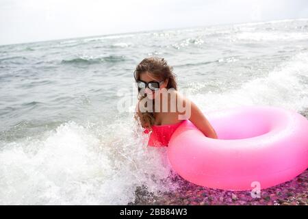 Junge Frau sitzt an der Küste oder am Meer in Wellen mit rosafarbener Luftmatratze in der Hand und genießt. Urlaubs- oder Reisezeit. Helle, farbenfrohe Luftmatratze a Stockfoto