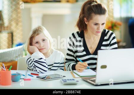 Langweilige Kinder erwecken Aufmerksamkeit, während die Mutter im Büro im modernen Wohnzimmer an sonnigen Tagen arbeitet. Stockfoto