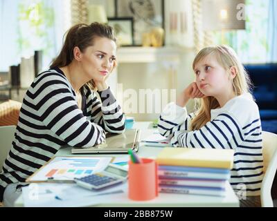 Traurige junge Mutter und Kind in gestreiften Pullovern im Heimbüro im modernen Haus am sonnigen Tag beim Arbeiten und bei Hausaufgaben einander angucken. Stockfoto