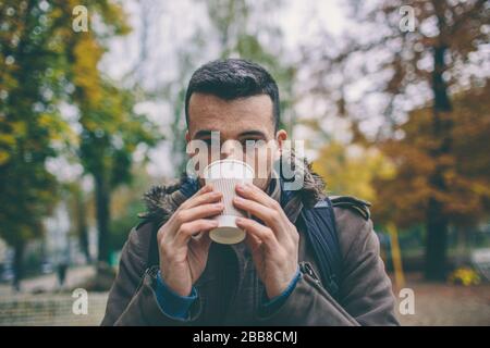 Mann im Mantel, der eine Tasse Kaffee Latte mit Milch hält. Einsame Frau steht auf einer verschneiten Herbststraße im Park. Stockfoto