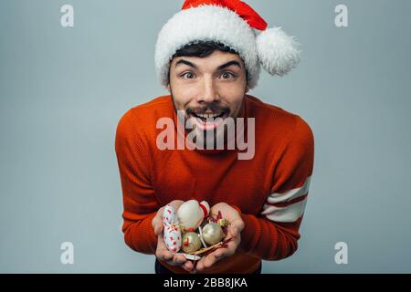 Junger bärtiger Mann isoliert über den Hintergrund. Aufgeregter positiver Kerl hält Dekorationsspielzeug für weihnachtsbaum in den Händen. Tragen Sie Santa's Hut und rote Feiertage c Stockfoto