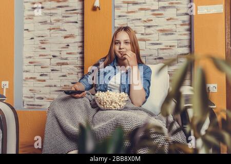 Entspanntes Mädchen im Teenager-Alter, das Fernsehen und Zapping mit Fernbedienung verfolgt und Popcorn zu Hause auf einem bequemen Sessel isst. Stockfoto