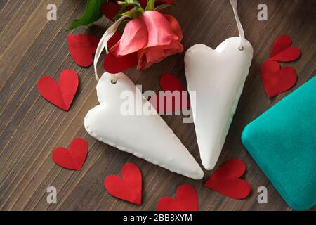 Zwei weiße Herzen aus Porzellan auf dem Holztexturtisch mit pinkfarbener Blume, blauem Kasten und vielen roten Papierherzen. Hochzeits- oder Liebeszeichen. Flaches Lay. Stockfoto