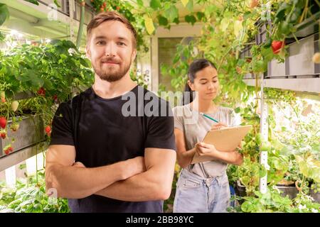 Portrait eines lächelnden, selbstbewussten jungen Mannes mit gekreuzten Armen, der mit Frau in der Baumschule arbeitet Stockfoto