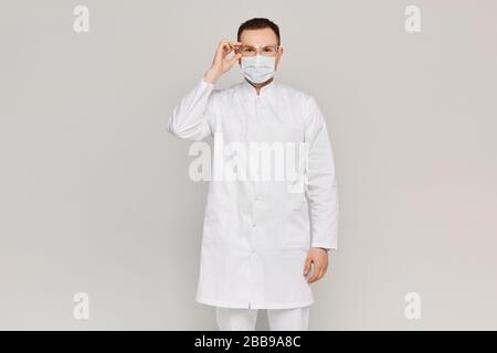 Gut aussehender Chirurg oder Arzt, der in einer Gesichtsmaske und einer Schutzbrille auf grauem Hintergrund posiert. Konzept des Gesundheitswesens. Schutz vor Grippe oder Virus Stockfoto