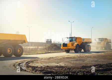 Viele große Gelenkwagen, die am sonnigen Tag mit blauem Himmel auf der neuen Straßenbaustelle fahren, sind mit gelben Müllhalden befahren Stockfoto