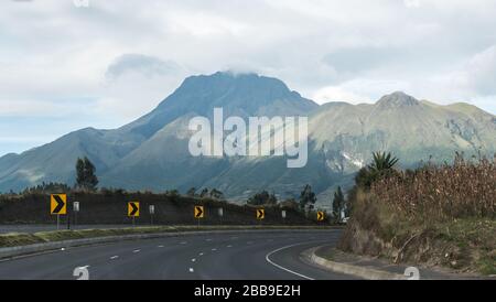 Große Pfeilschilder zeigen den Weg einer scharfen Wendung auf einer Autobahn in Ecuador mit dem großen inaktiven Vulkan Imbabura im Hintergrund. Stockfoto