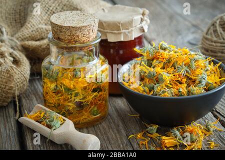 Flaschen Kalendula-Infusion oder Öl, gesunde Marigold-Blumen in Schüssel und Calendula-Salve auf Holztisch. Pflanzenheilkunde. Stockfoto
