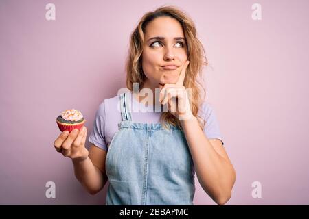 Junge schöne blonde Frau Eatimg Schokoladenkuchen über isoliertem rosa Hintergrund ernsthaftes Gesicht Denken über Frage, sehr verwirrte Idee Stockfoto