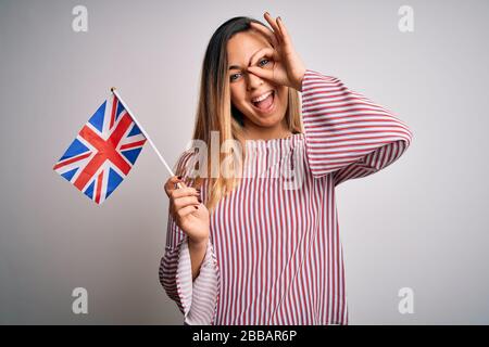 Junge, schöne blonde Frau mit blauen Augen, die die flagge des vereinigten Königreichs hält, mit einem fröhlichen Gesicht, das lächelt, und einem guten Schild mit der Hand auf dem Auge, das durch die Finger blickt Stockfoto