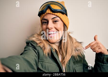 Junge Brunette-Skifahrerin, die Schneekleidung und eine Skibrille trägt, macht selfie per Kamera mit überraschtem Gesicht, das mit dem Finger auf sich zeigt Stockfoto