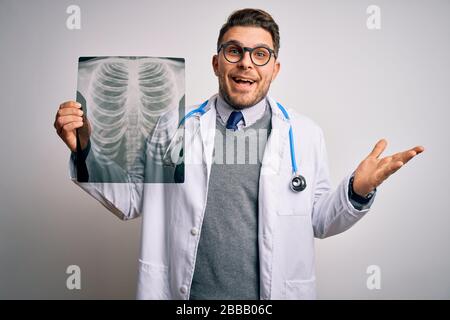 Junger Arzt, der einen medizinischen Mantel trägt, der die Radiographie des Brustkorbs über einem isolierten Hintergrund betrachtet, sehr glücklich und aufgeregt, der Siegerausdruck feiert Stockfoto