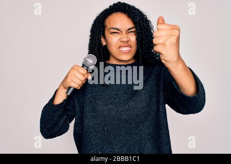 Junge afroamerikanische Curly Singer-Frau, die mit Mikrofon über weißem Hintergrund singt, ärgerte sich und frustrierte Schreie mit Wut, Wahnsinn und Geschrei Stockfoto