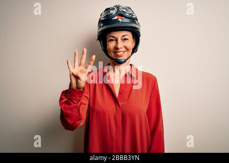 Motorradfahrerin im mittleren Alter, die einen Motorradhelm über einem isolierten weißen Hintergrund trägt, der mit den Fingern Nummer vier zeigt und nach oben zeigt, während sie lächelt Stockfoto