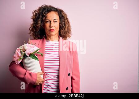 Schöne Frau im mittleren Alter, die Vase mit Blumen über isoliertem rosafarbenem Hintergrund hält, mit einem selbstbewussten Ausdruck auf einem klugen Gesicht, das ernsthaft denkt Stockfoto