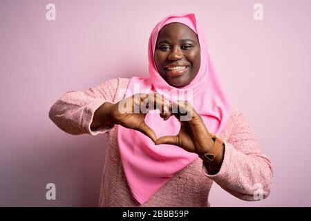 Junge afroamerikanische und große Frau mit muslimischem Hijab über isoliertem rosafarbenem Hintergrund, die in Liebe lächelnd mit Händen die Form eines Herzsymbols macht. Romanti Stockfoto