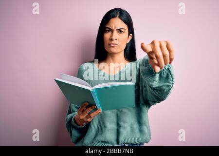 Junge schöne hispanische Studentin liest ein Buch über rosa isoliertem Hintergrund und zeigt mit dem Finger auf die Kamera und auf Sie, Handzeichen, positiv Stockfoto