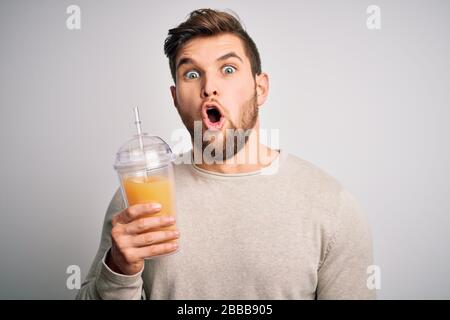 Junger blonder Mann mit Bart und blauen Augen, der einen gesunden orangefarbenen Smoothie trinkt, erschreckt mit einem überraschenden Gesicht, Angst und Begeisterung mit Angst Expressio Stockfoto