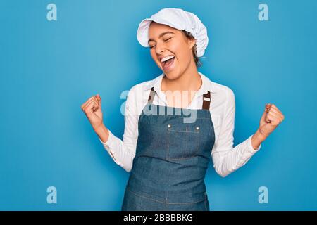 Junge, schöne blonde Bäckerin mit blauen Augen, die mit Schürze und Kappe kocht, sehr glücklich und aufgeregt, wenn sie Gewinnergeste mit erhobenen Armen und lächelnden Armen macht Stockfoto