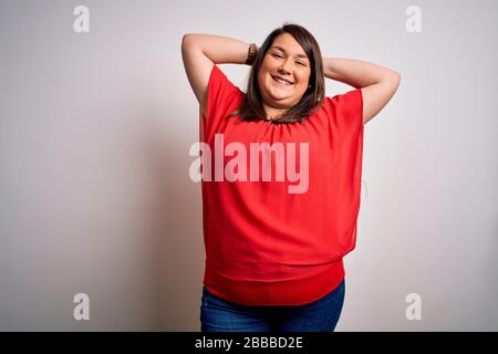 Schöne Brunette und große Frau mit legeren roten T-Shirts über isoliertem weißem Hintergrund entspannend und streckt, Arme und Hände hinter Kopf und n Stockfoto