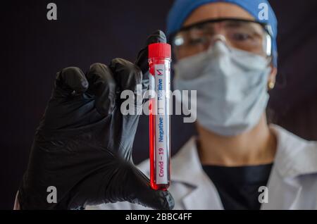 Ein weiblicher Arzt, der eine chirurgische Maske und eine Schutzbrille trägt, zeigt ein Covid-19-Bluttestrohr. Positives Ergebnis des Bluttestrohrs von Coronavirus. Stockfoto