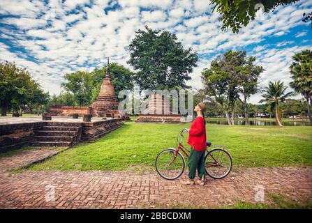Frau im roten t-Shirt mit Fahrrad, Blick auf alten verfallenen buddhistischen Tempel in Geschichtspark Sukhothai, Thailand Stockfoto