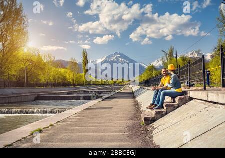 Glückliches Paar in Blau und Gelb Kostüme auf der Treppe in der Nähe des Flusses und die schneebedeckten Berge im Hintergrund in Almaty, Kasachstan sitzen Stockfoto