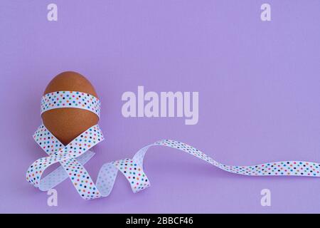 Ei steht auf Becher, auf weichem violettem Hintergrund. Osterei mit festlichem Band gefesselt Stockfoto