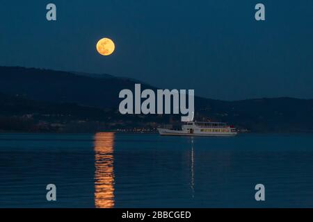 Vollmond steigt über einige Hügel auf einem See und reflektiert perfekt auf Wasser in der Nähe eines eintreffenden Bootes Stockfoto