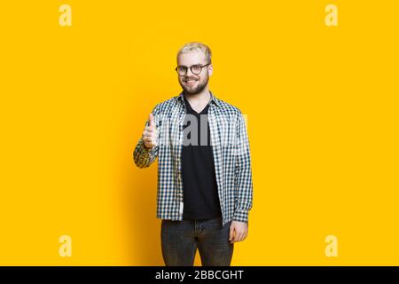 Kaukasischer bärtiger Mann mit Brille und blondem Haar trägt das Annäherungszeichen in einer gelben Wand Stockfoto