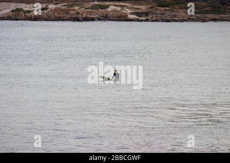 Kapstadt, Südafrika. März 2020. Ein einsamer Surfer sitzt auf seinem Brett, während er wartet, bis Wellen auf einem ruhigen Ozean hereinkommen. Stockfoto