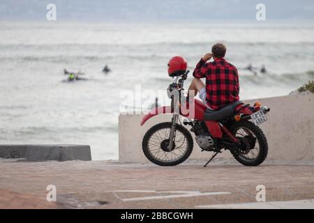 Muizenberg, Südafrika. März 2020. Ein Kerl auf einem Fahrrad, das rot trägt, sitzt auf seinem Fahrrad und entspannt sich, während er den Ozean und Surfer in der Ferne beobachtet. Stockfoto