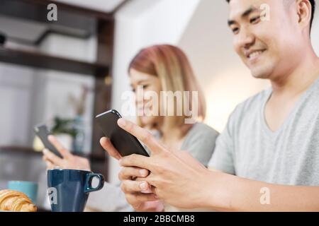 Glückliches asiatisches Paar in Schlafanzug sitzt am Tisch in der Küche und sieht Video auf dem mobilen Togerther zu Hause am Morgen und mit Müsli und Cornflakes Frühstück.Focus Stockfoto