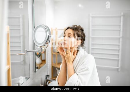 Junge Frau im bademantel, die sich um ihre Haut kümmert und auf den Spiegel im Bad schaut. Gesichtshautpflege und Wellness-Konzept Stockfoto