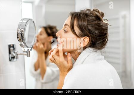 Junge Frau im bademantel, die sich um ihre Haut kümmert, auf den Spiegel schaut und sich um Falten im Bad sorgt. Gesichtshautpflegekonzept Stockfoto