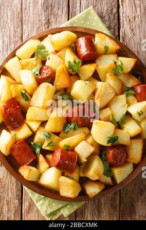 Bratkartoffeln im Landhausstil mit Würstchen und Kräutern liegen in einem Teller auf dem Tisch. Vertikale Draufsicht von oben Stockfoto