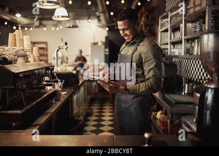 Fröhlicher junger afro-amerikanischer Café-Besitzer, der schwarze gestreifte Schürze mit digitalem Tablet trägt Stockfoto