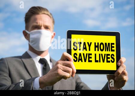 Geschäftsmann, der eine COVID-19-Gesichtsmaske trägt, die ein Tablet mit einem "Stay Home"-Tablet hält, rettet Lebensnachrichten Stockfoto