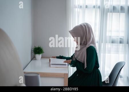 Hijab-Frauen, die auf dem Stuhl sitzen, studieren und lesen das heilige Buch Al-quran im Schlafzimmer Stockfoto
