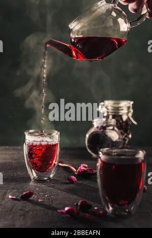 Verarbeiten Sie roten Karkade-Tee, Rosentee, Tasse frisch gebrühten Fruchttee, dunkles Moody. Heißes Wasser wird aus dem Wasserkocher in eine flass Tasse gefüllt Stockfoto